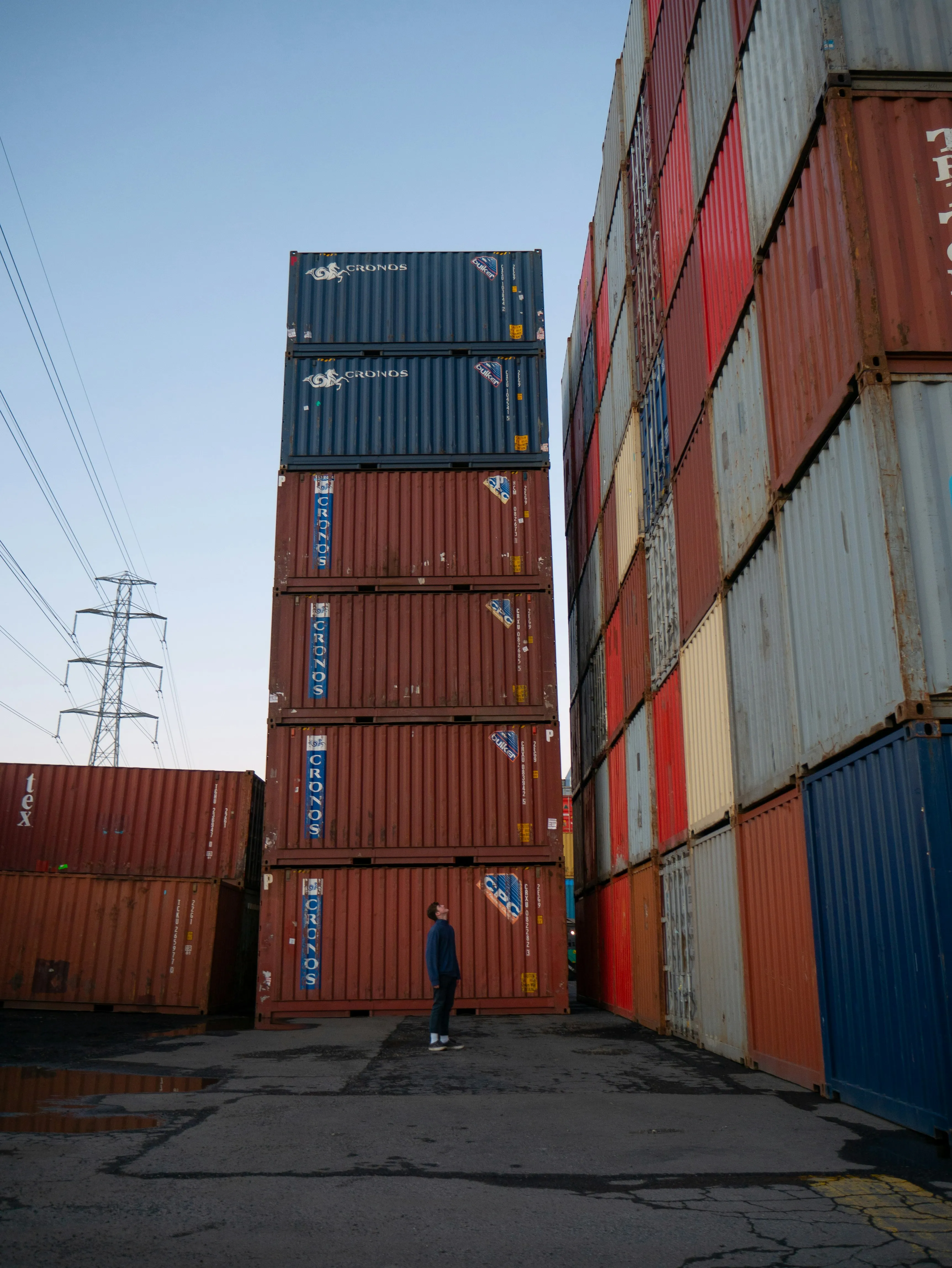 A person looking up at a tall stack of shipping containers in a container yard
